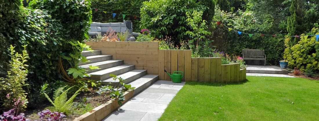 Woodland garden on a sloping plot with an upper terraced seating area, wide sandstone steps, sleeper retaining walls and a lower level lawn.