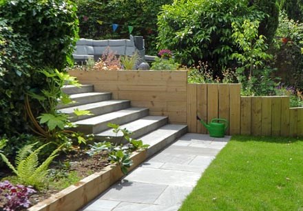 Woodland garden on a sloping plot with an upper terraced seating area, wide sandstone steps, sleeper retaining walls and a lower level lawn.